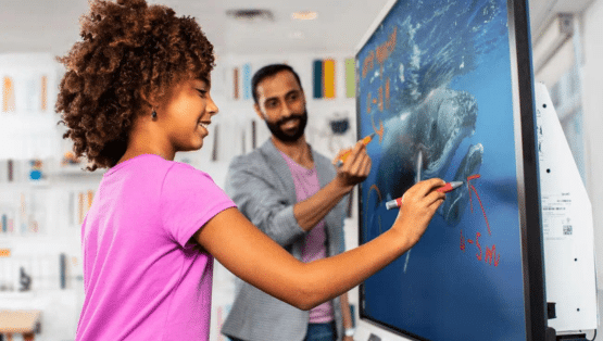Een schoolmeisje staat voor een SMART Board samen met haar leraar en maakt gebruik van SMART software. Ze heeft een roze shirt aan.
