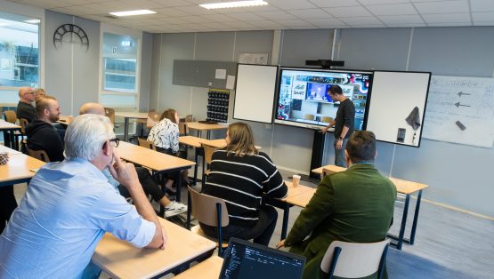 Digibord in klaslokaal met interactieve functies, geschikt voor aanbestedingen en marktconsultatie. Een docent staat voor het bord en geïnteresseerden luisteren aandachtig naar de docent en kijken naar het digibord.