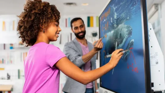 Een leerling staat voor een digibord (touchscreen) samen met haar leraar en maakt gebruik van SMART software. Ze heeft een roze shirt aan en tekent op het interactieve scherm. | Voordelen van een digibord.